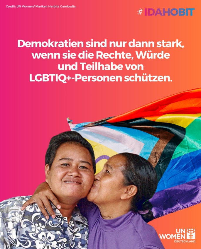Frau küsst ihre Partnerin auf die Wange, dahinter eine Pride Flagge