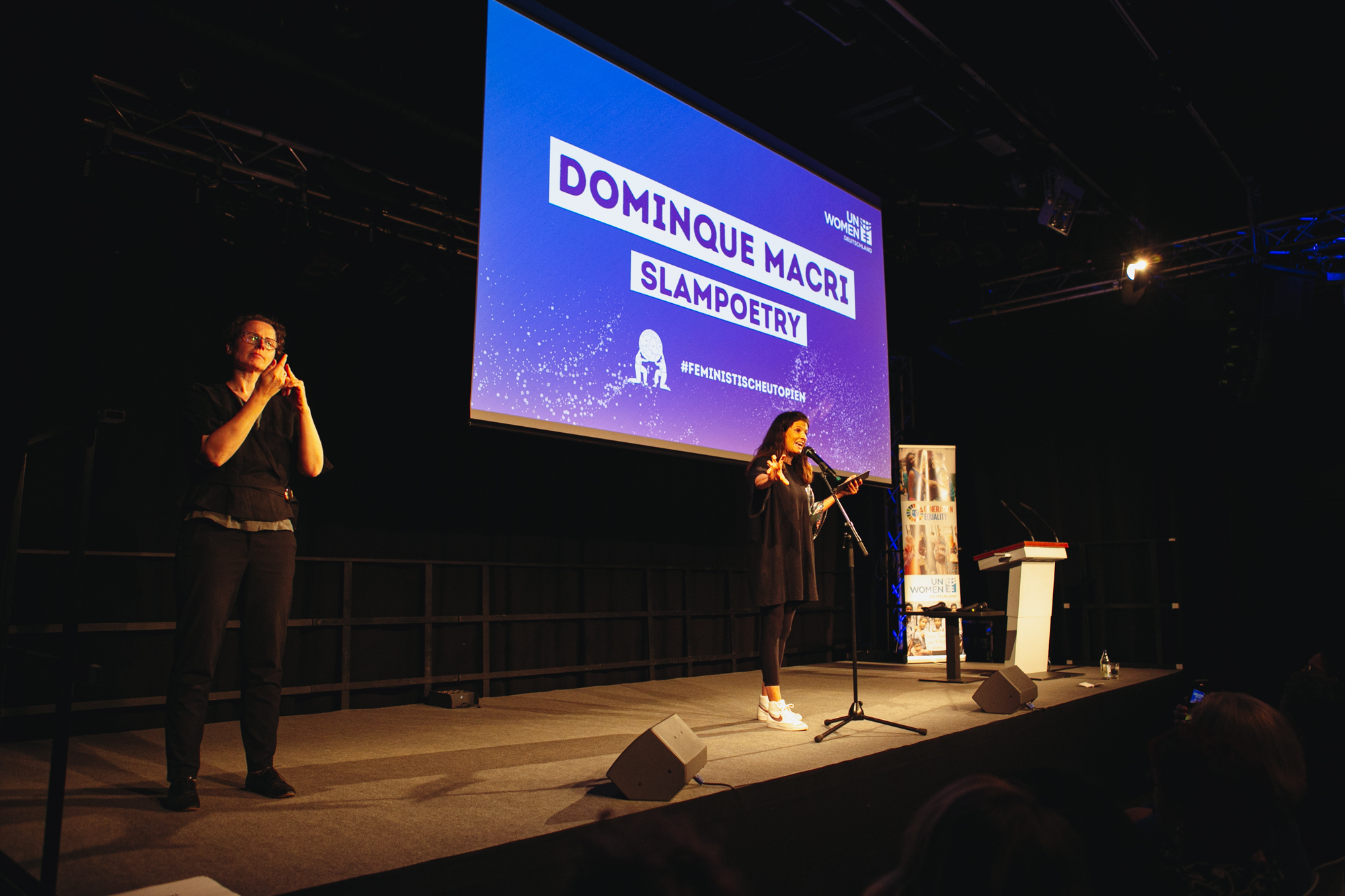 Dominique Macri stellt ihre lyrische Zusammenfassung auf der Bühne vor. Die Gebärdendolmetscherin steht links auf der Bühne und übersetzt die Inhalte von der Slampoetin.