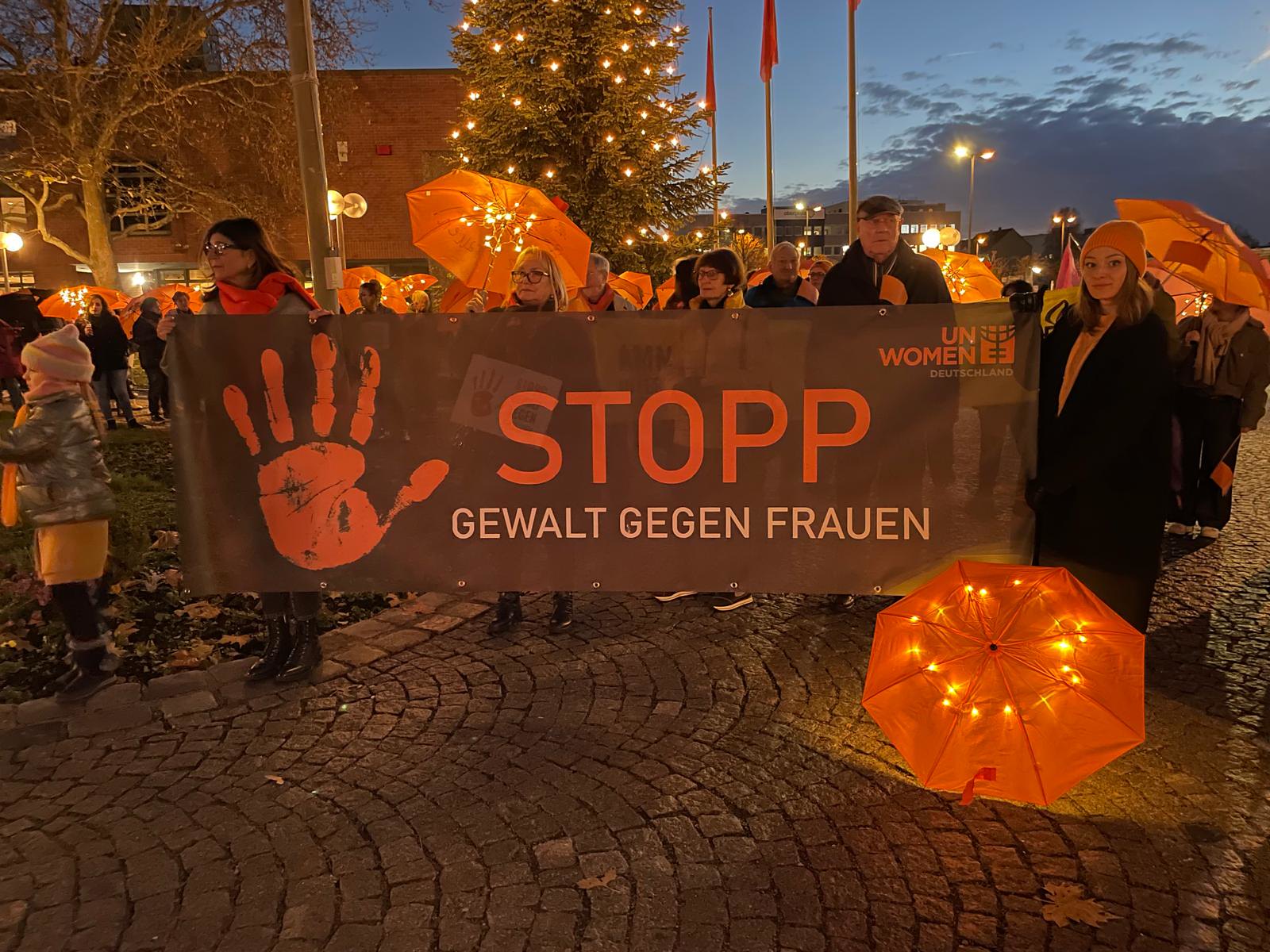 Menschen versammeln sich hinter Orange The World Plakat - Stopp Gewalt gegen Frauen.