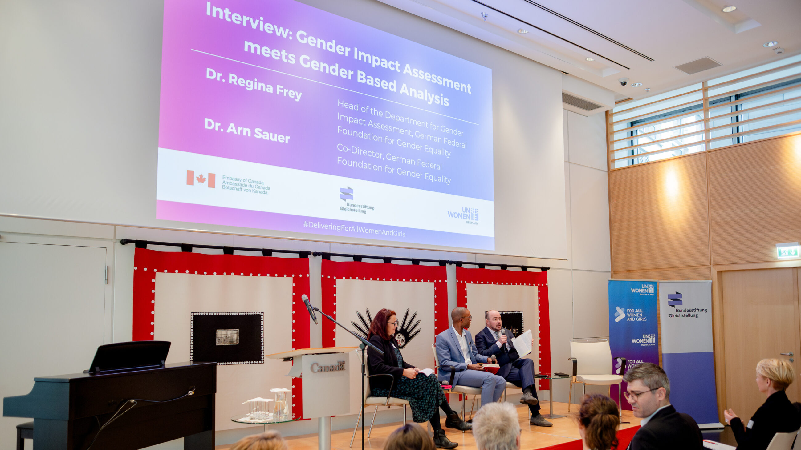 Dr. Regina Frey and Dr. Arn Sauer on stage at the international conference. They are being interviewed by the moderator Jesse George who is sitting between them.