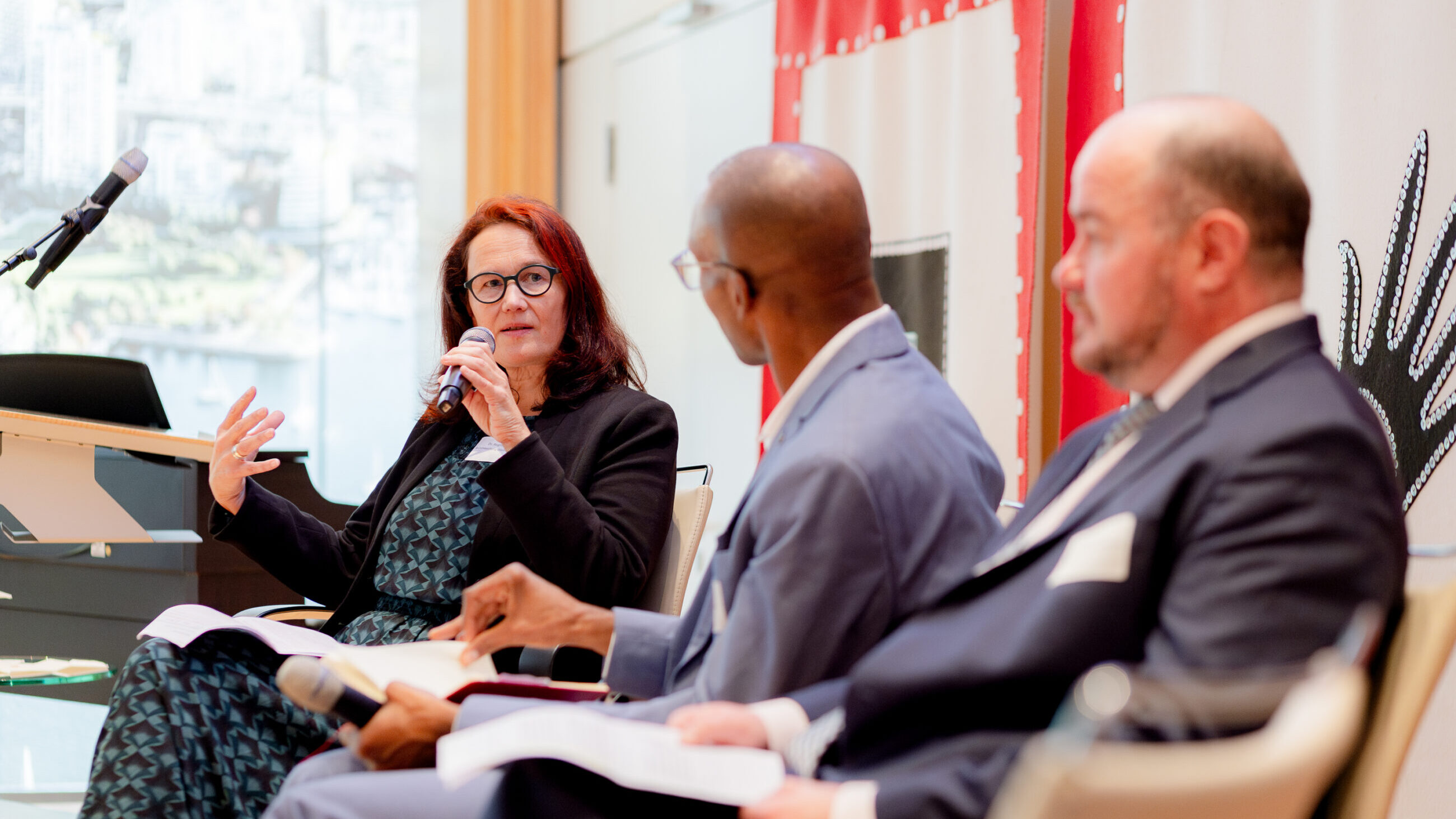 Dr. Regina Frey and Dr. Arn Sauer on stage at the international conference. They are being interviewed by the moderator Jesse George who is sitting between them.