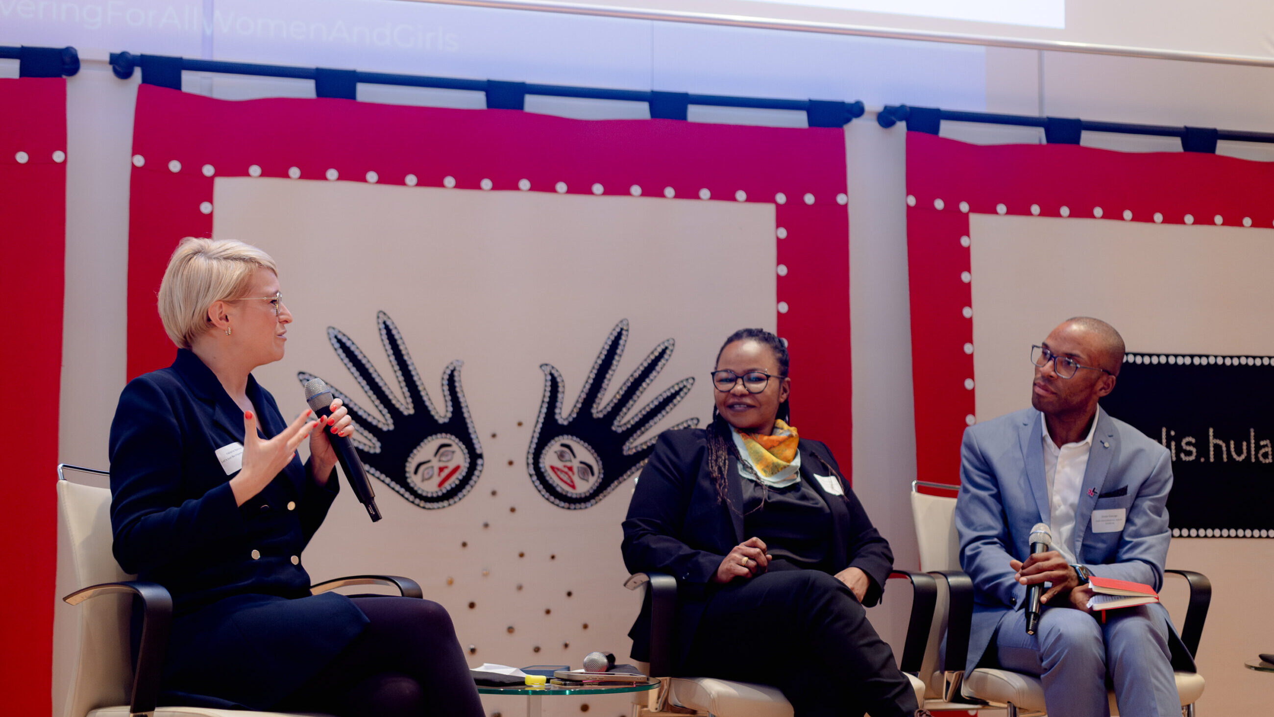 A participant from the audience is speaking on stage next to Mai Ali Shatta and Jesse George.