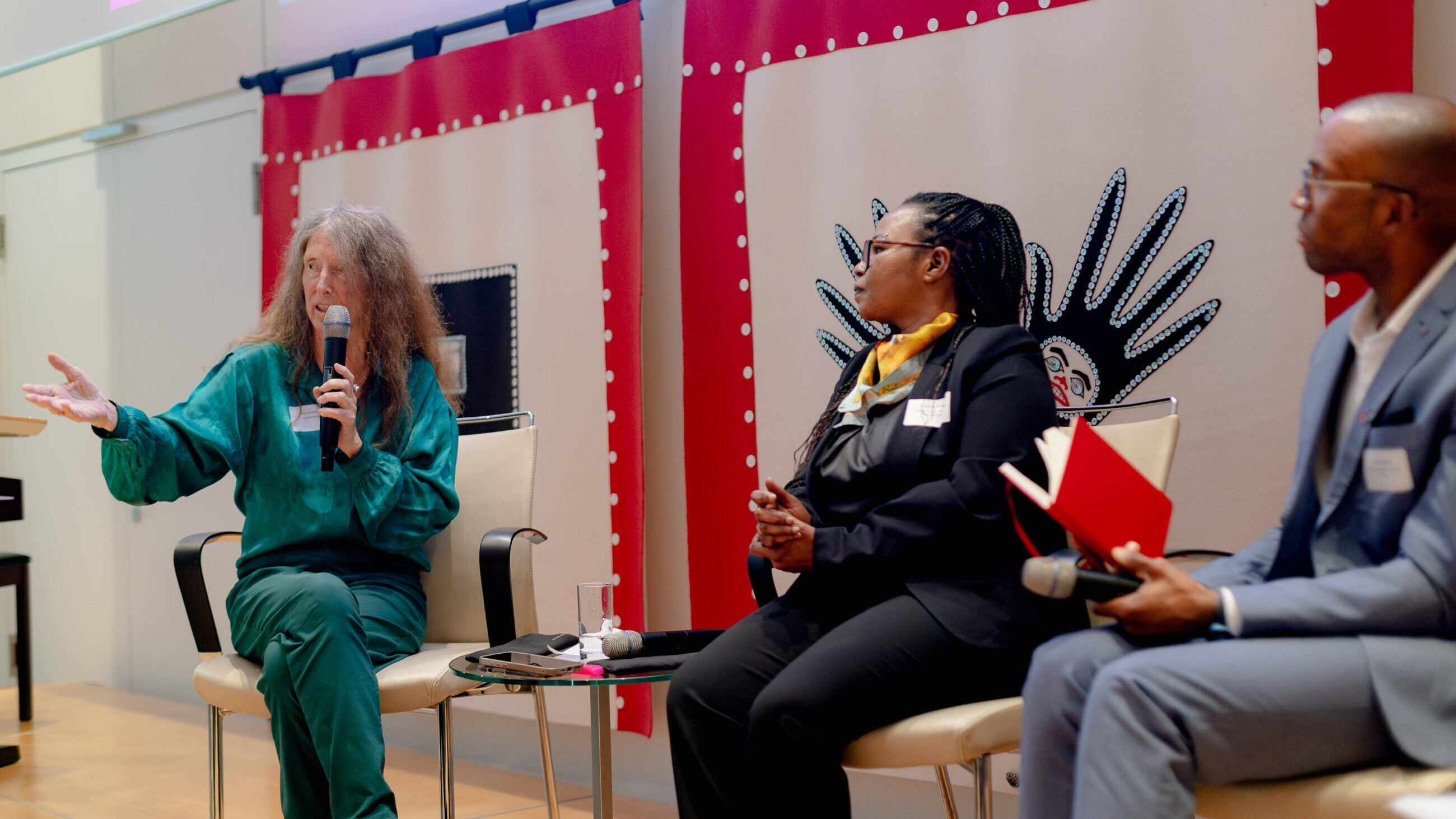 A participant from the audience is speaking on stage next to Mai Ali Shatta and Jesse George.