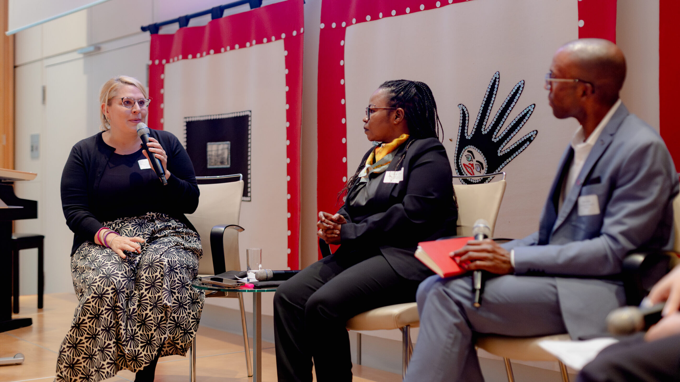 A participant from the audience is speaking on stage next to Mai Ali Shatta and Jesse George.