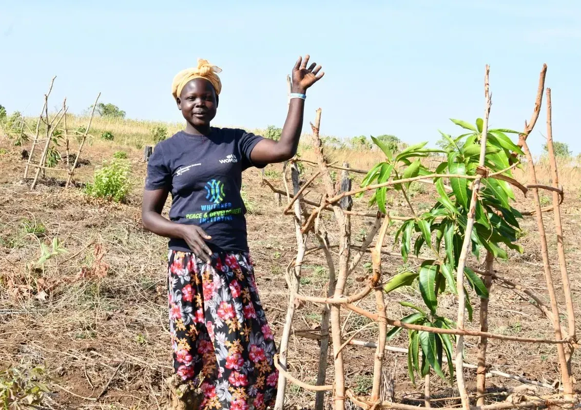 Frau winkt in die Kamera. Sie steht inmitten einer Farm neben jungen Mango Baum