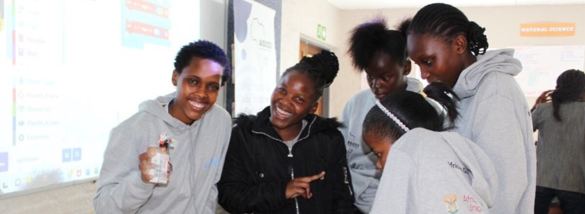 Eine Gruppe Mädchen steht vor einem Laptop. Zwei lächeln in die Kamera. African Girls Can Code Projekt - Pekinger Erklärung und Aktionsplattform - Bejing+30